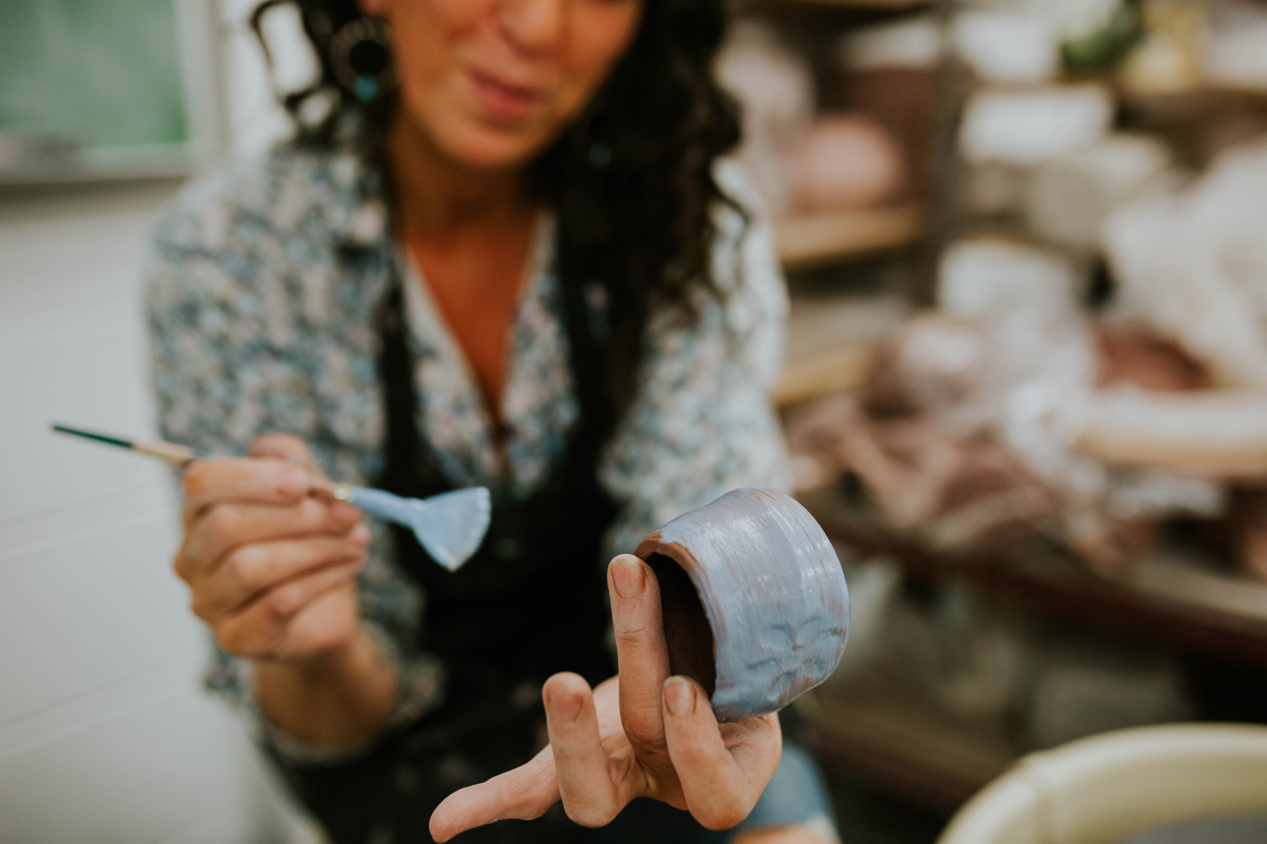 A woman painting a light blue glaze onto a red clay mug to then fire in a kiln to create a piece of ceramics.