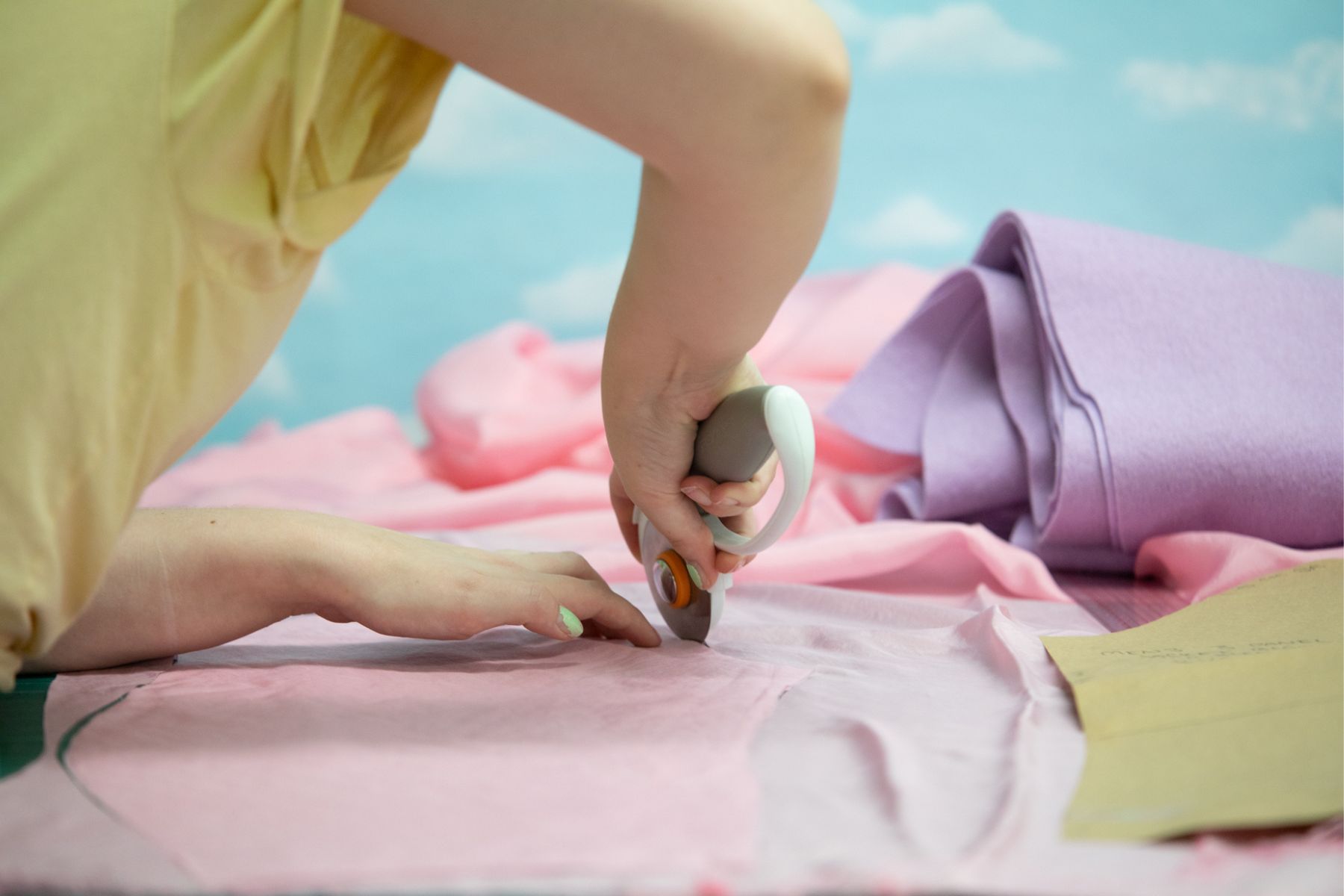 A person cuts a piece of pink fabric at the Honeybee Folk School. The wallpaper resembles clouds in the background.