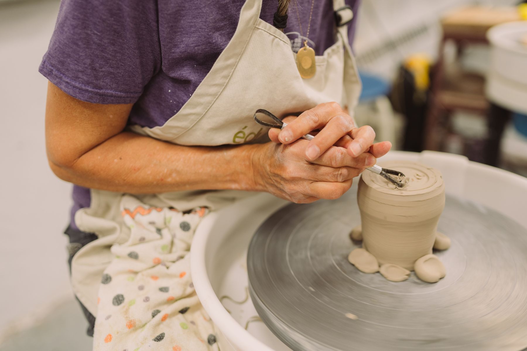 A Honeybee Folk School workshop participant shaves down a clay cylinder on a pottery wheel. It is a close-up of her hands.